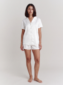 A woman modeling a white short-sleeve button-up shirt and matching white shorts pajama set against a plain light gray studio background.
