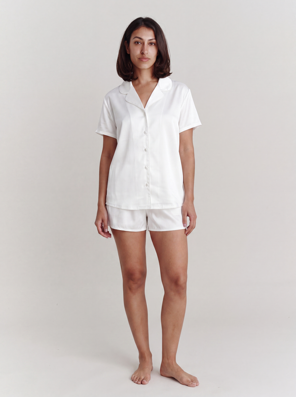 A woman modeling a white short-sleeve button-up shirt and matching white shorts pajama set against a plain light gray studio background.