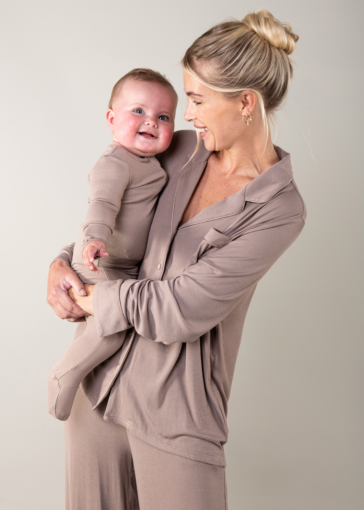 Woman holding a baby and both wearing matching beige outfits against a plain background