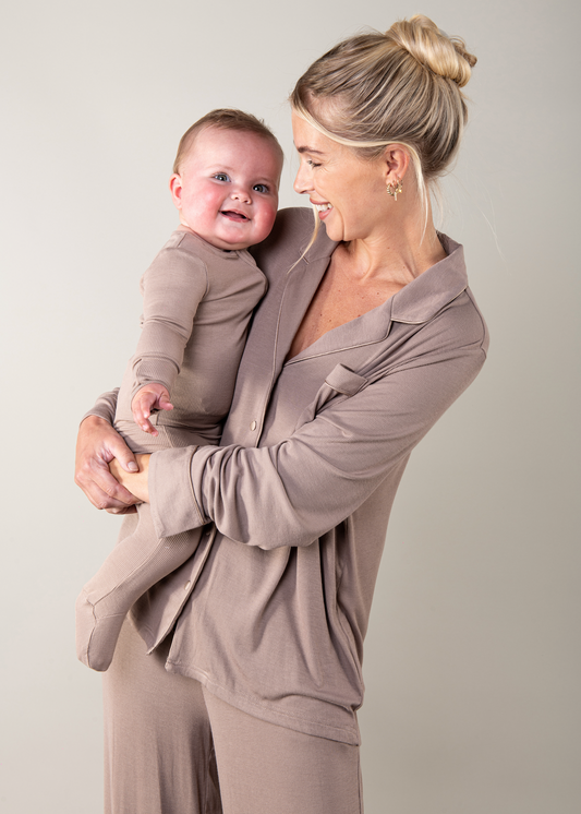 Woman holding a baby and both wearing matching beige outfits against a plain background